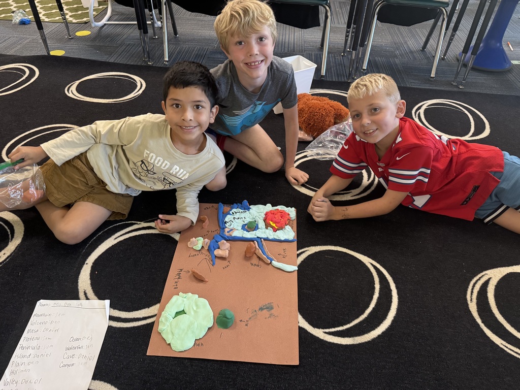 Group of boys sitting on carpet with a clipboard and a piece of paper visible and clay as well made into animal landforms.