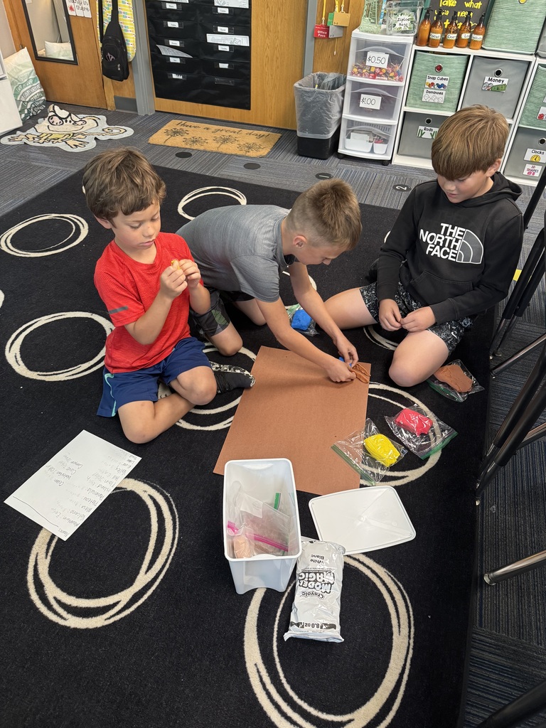 Group of boys sitting on carpet with a clipboard and a piece of paper visible and clay as well.