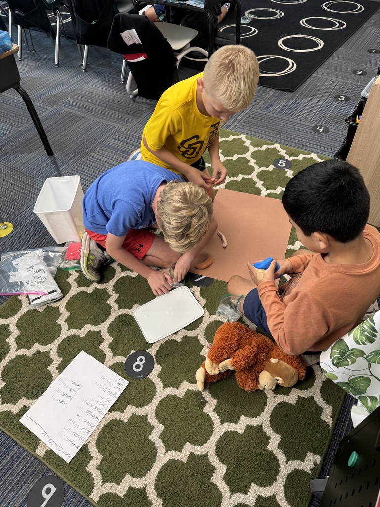 Three boys sitting around a counter with a clipboard and a piece of paper visible along with clay.