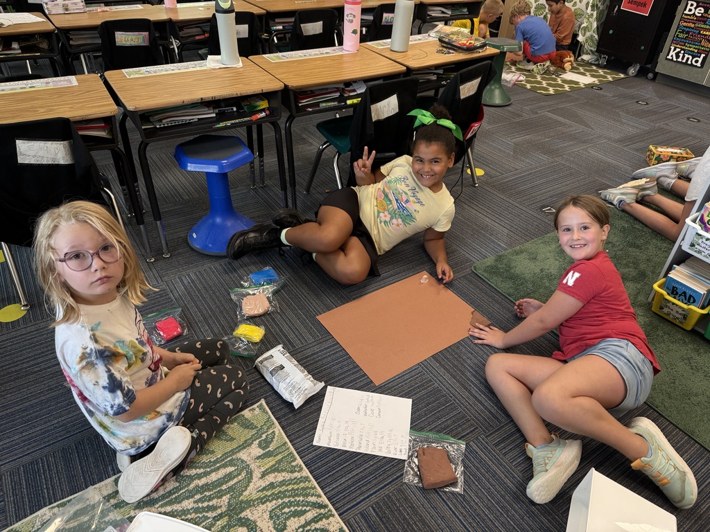Group of girls sitting on carpet with a clipboard and a piece of paper visible and clay as well.