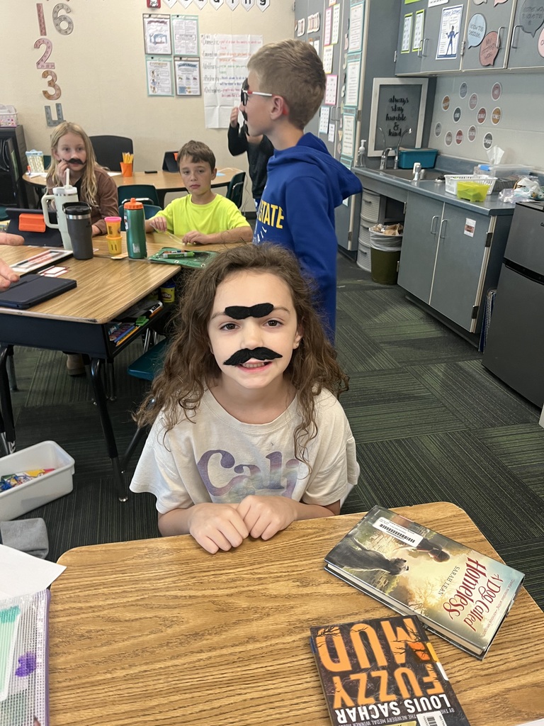 Girl sitting at a desk with books with a fake mustache and fake eyebrows.