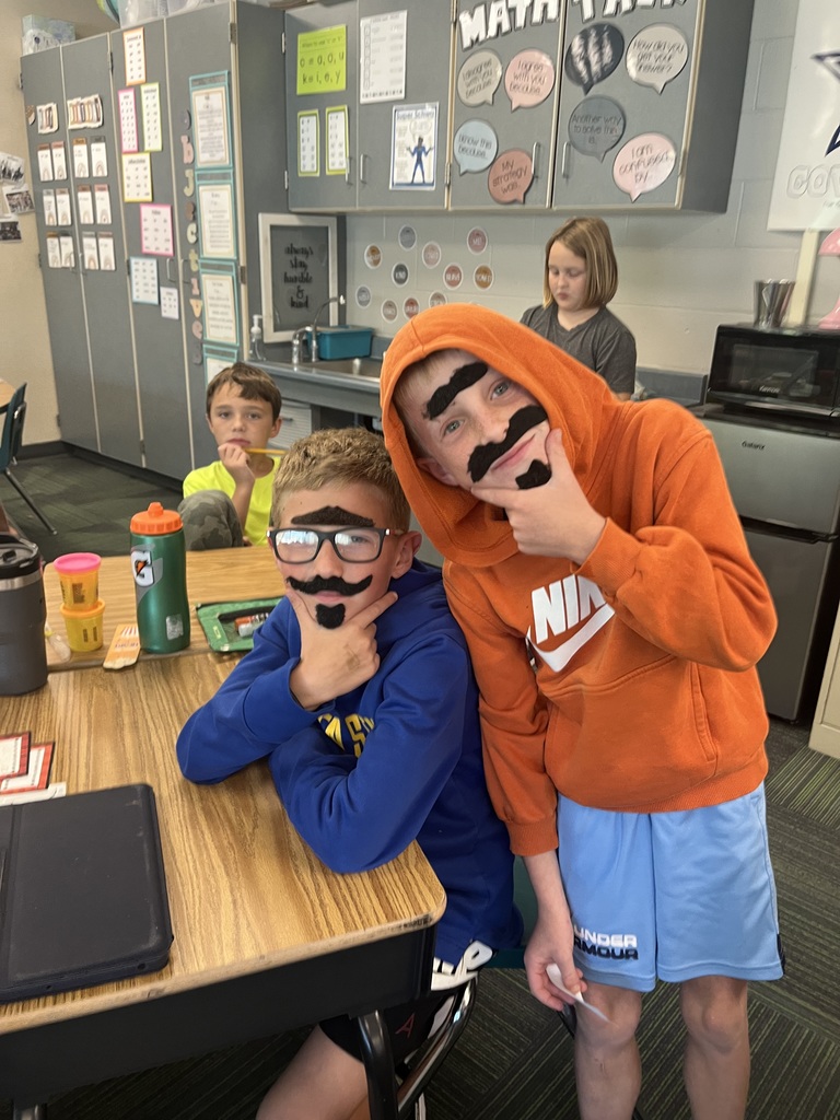 Two boys, one standing and one sitting at a desk, wearing fake mustaches and beards.