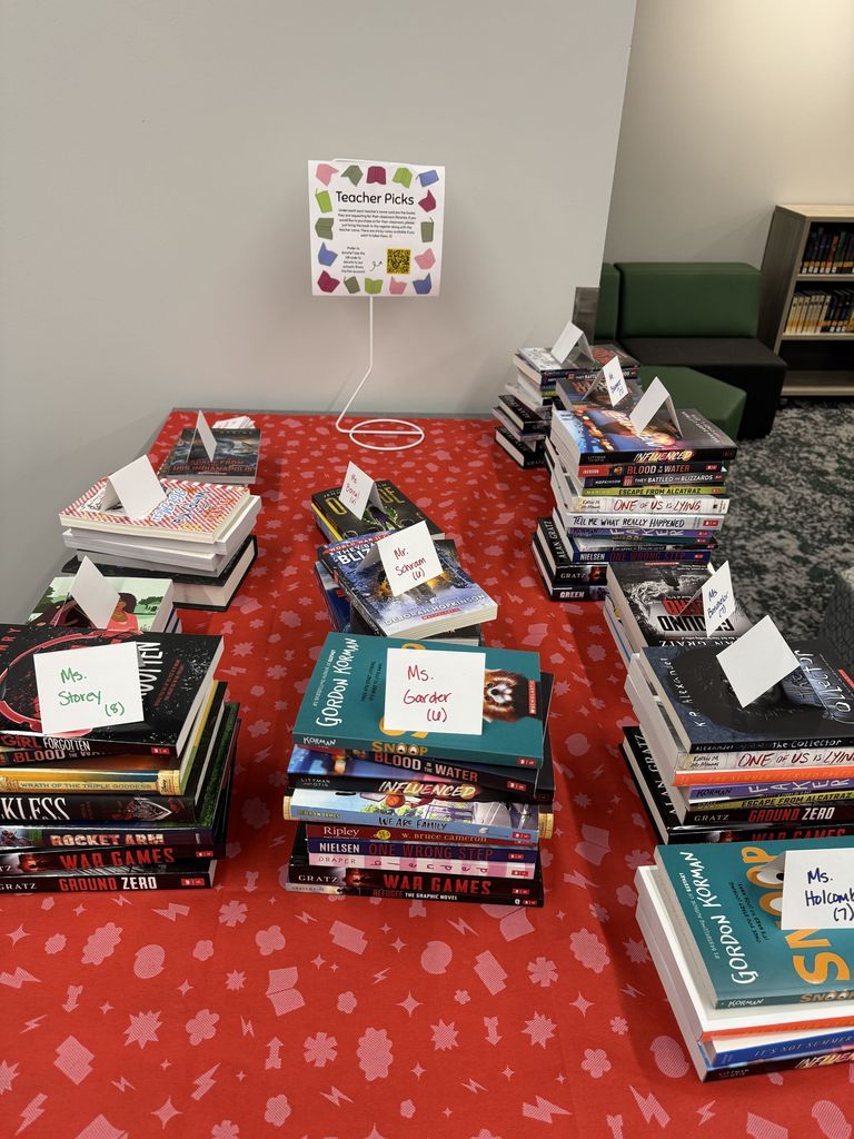 Stacks of books with teacher names on a table with a sign promoting teacher picks. 