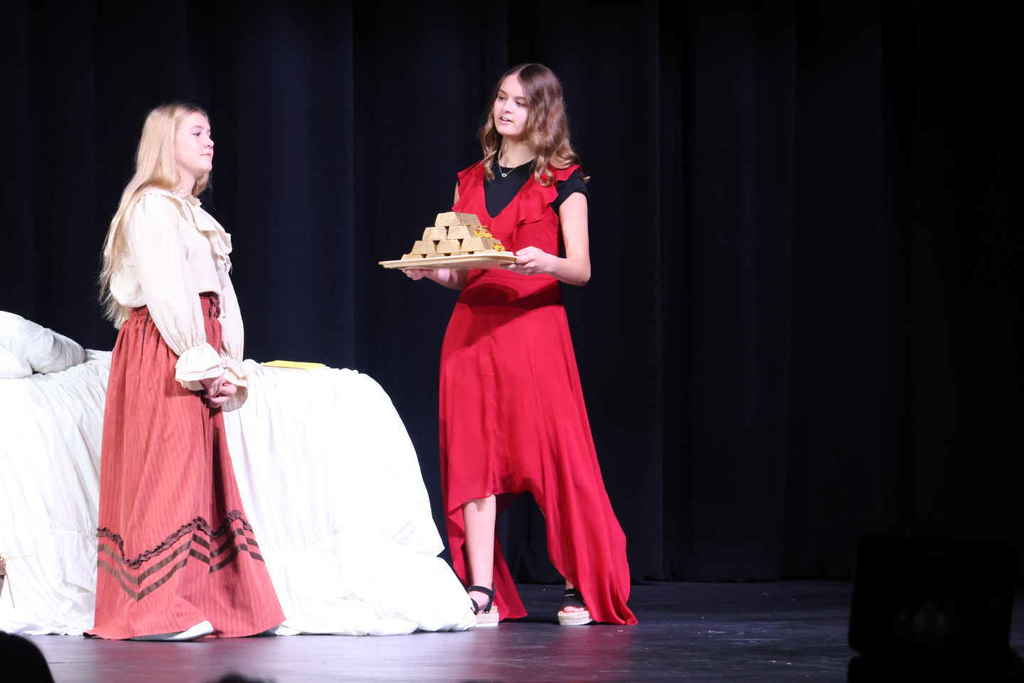 Two female students in the one act play, one holding a platter and the other standing next to a bed. 