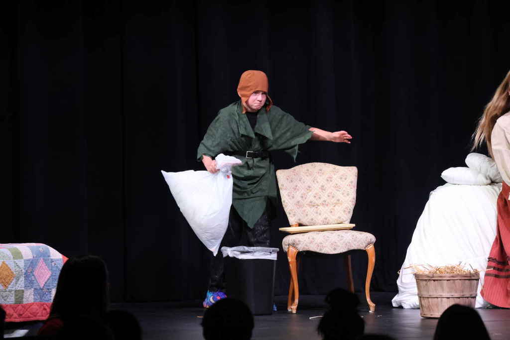 Student in the one act play, standing next to a chair and a trash can while holding a pillow case. 