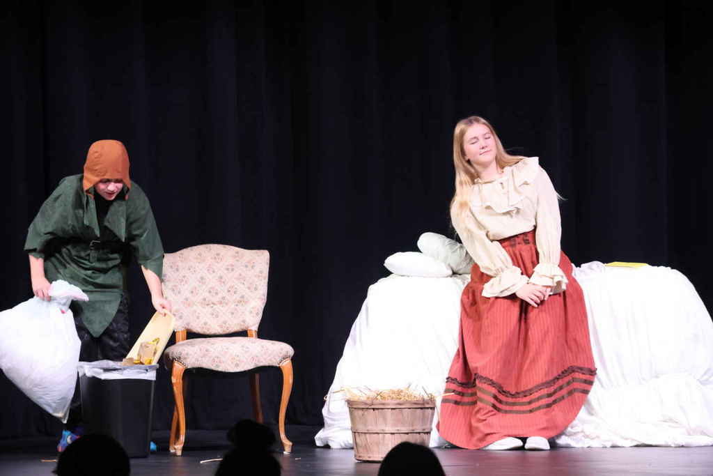 Students in the one act play. One student is emptying the trash and the other is sitting on a bed. 