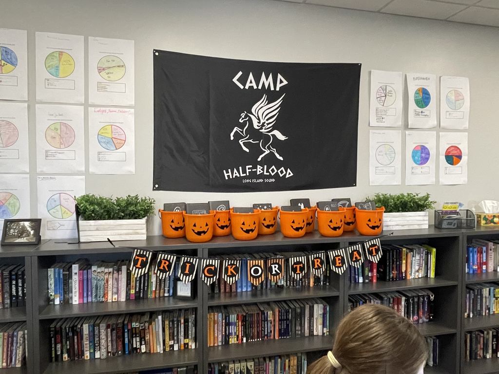 Pumpkin halloween buckets sit on a counter in a classroom. The pumpkins have books wrapped in black wrapping paper inside them with a banner in front that says Trick or Treat. 