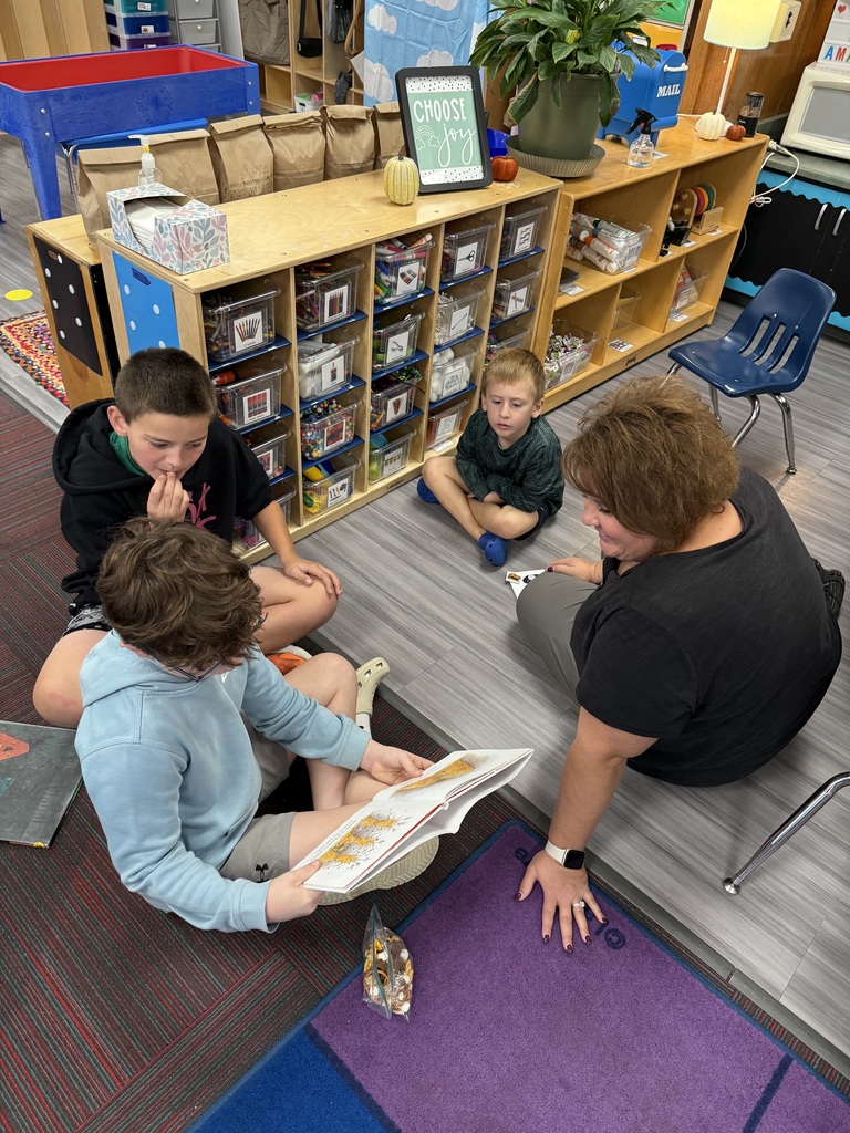 Three boys sitting on the floor reading a book together with an adult looking on.