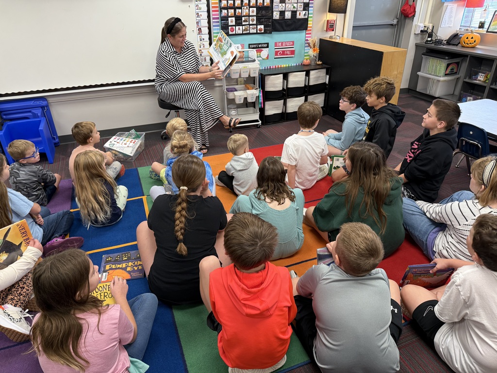 Teacher sitting at the front of the room reading a book to a group of students sitting on the floor.