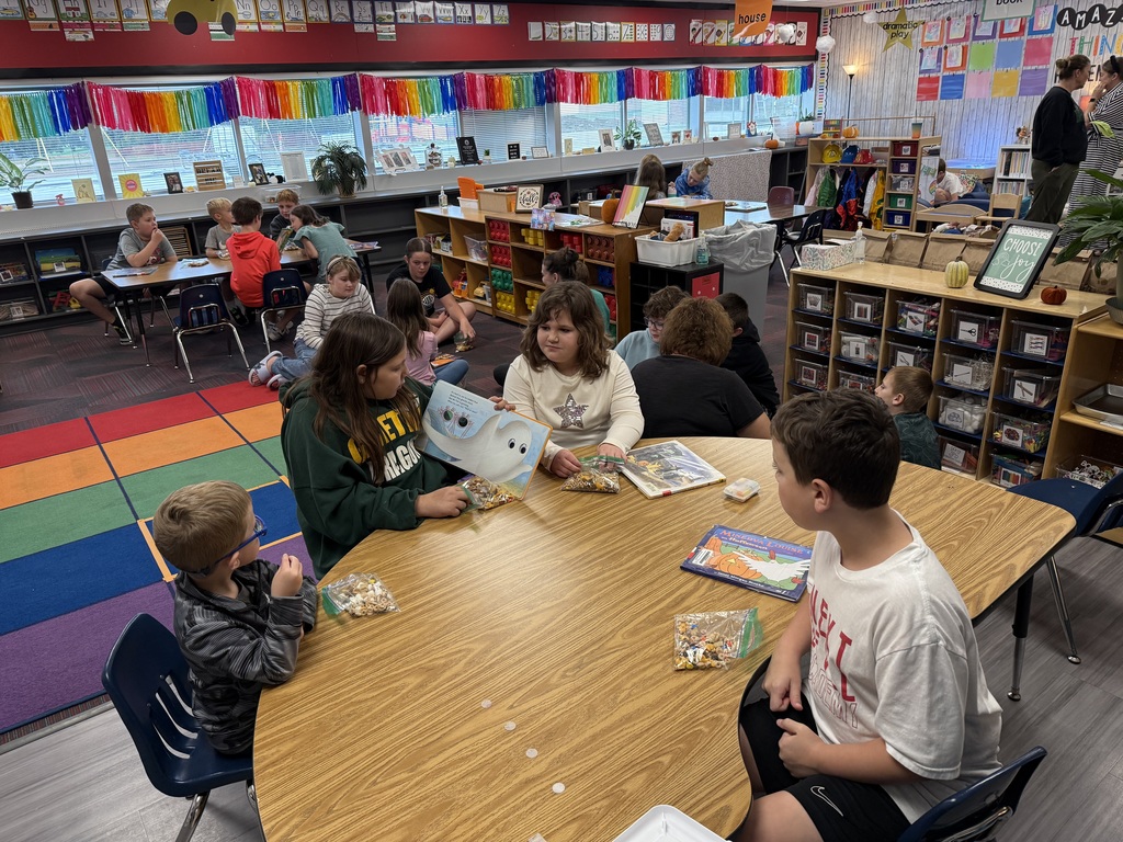 Three students sitting a kidney table reading a book to a yougner student with other students sitting in the background on the ground.