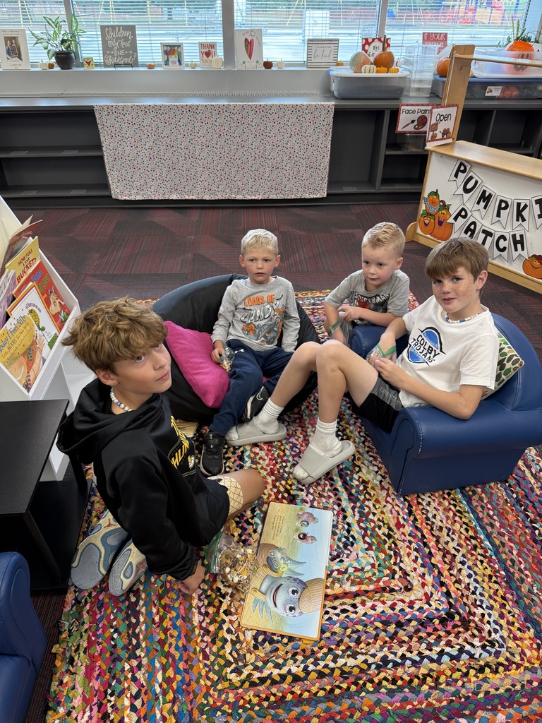 Two older boys sitting on a rug and chair reading a book to two younger students.