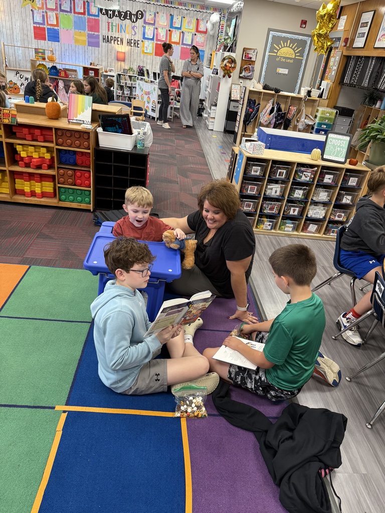 Two older boys sitting on colored carpet reading a story to a boy sitting at a small desk with an adult sitting next to them.