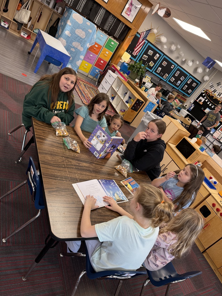 Group of girls sitting at a brown table reading three books while eating a snack.