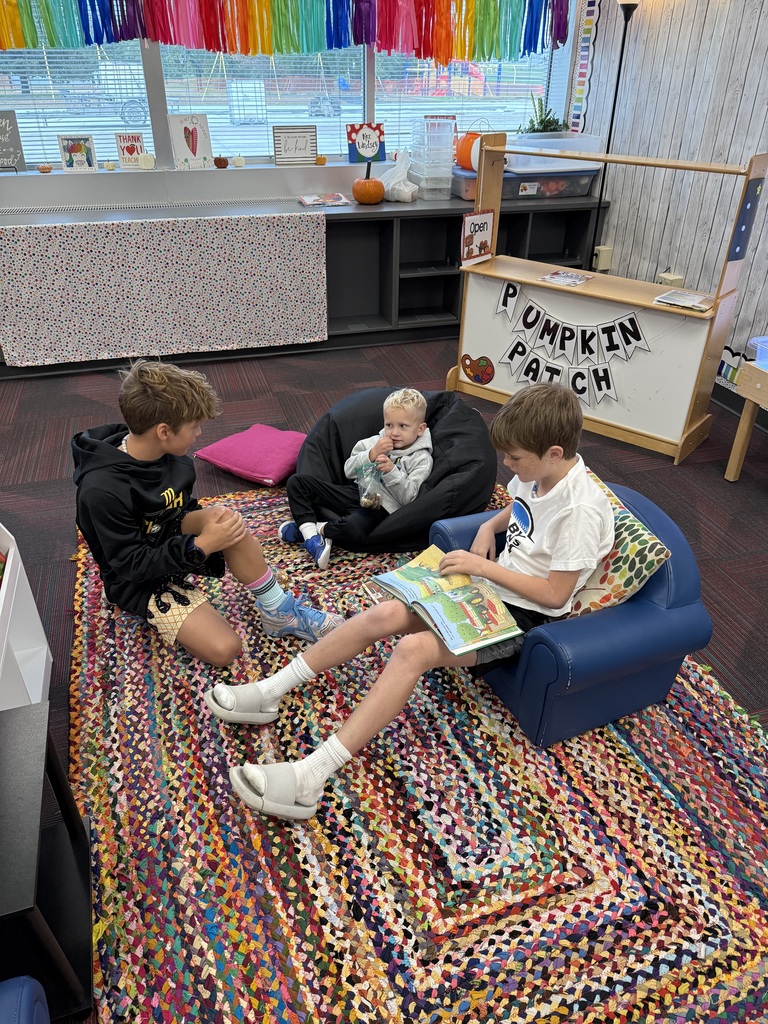 3 boys, one sitting on a rug and two sitting in comfy chairs reading a book.