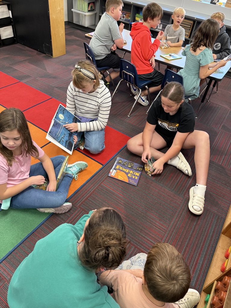 3 students sitting on the floor reading a book about pumpkins to a student with an adult sitting next to them.