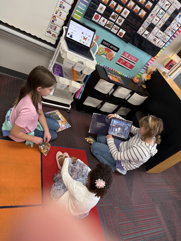 Two older girls sitting on the ground reading a book to a younger student who is sitting on the ground.