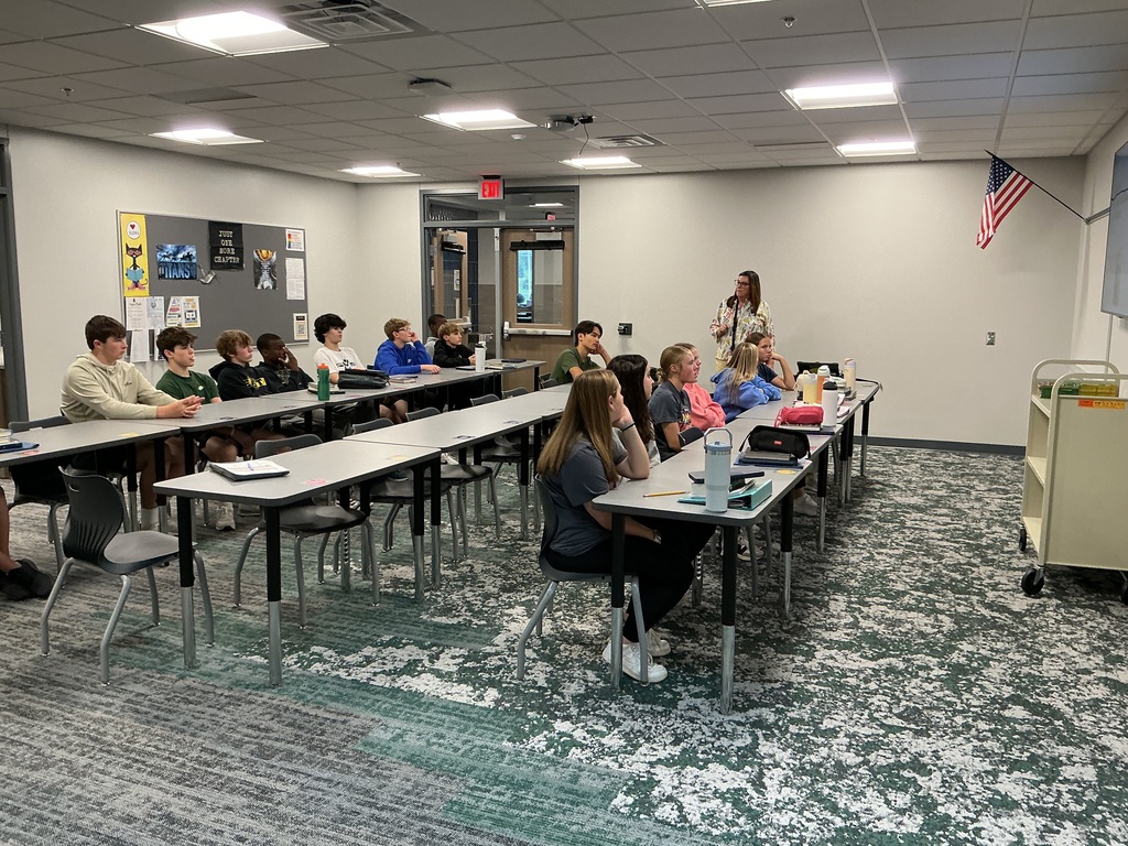 Photo of students sitting at tables listening to the teacher present.
