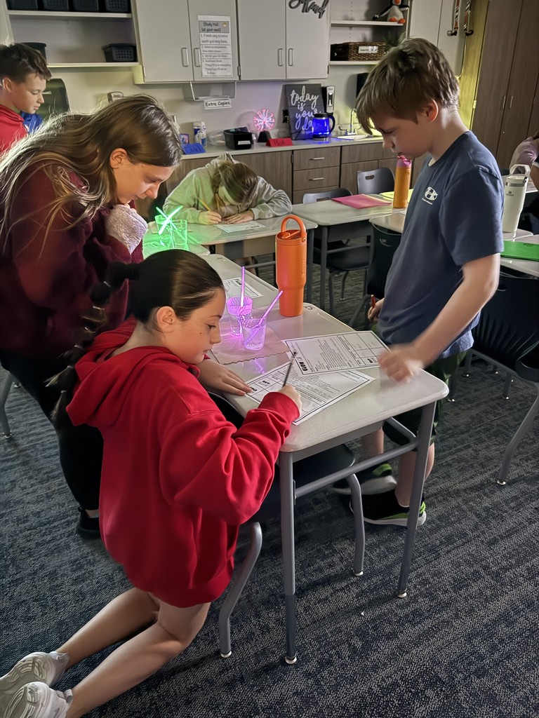 Students working at desks with a glowing item in a cup of liquid. They are taking notes. 