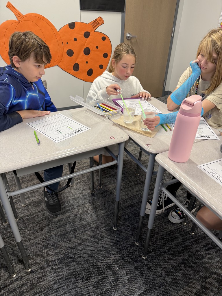Students preparing the glow lab activity. Three students sit at desks and are looking at a cup with glowing straws and taking notes. 
