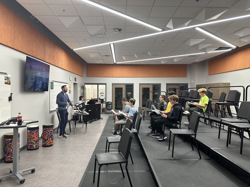 Six students sit in chairs on risers singing with a teacher in the front of the room and an accompanistat the piano. 
