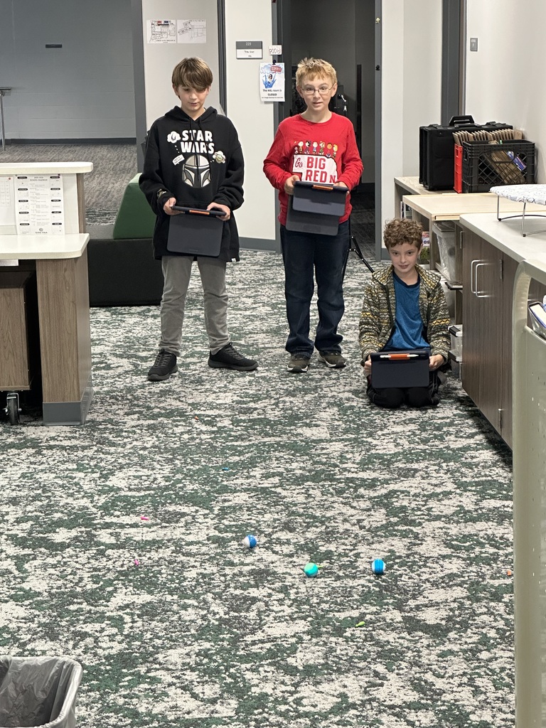 Three students are using iPads to drive robots on the carpet. 