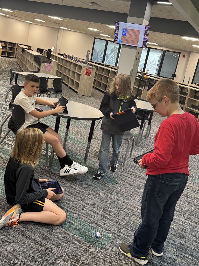 Four students are around a circular table with iPads. There are small robots on the floor. 