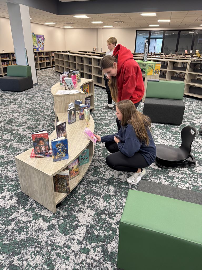 Three students are looking at books on a display case in a library. One is kneeling down. 