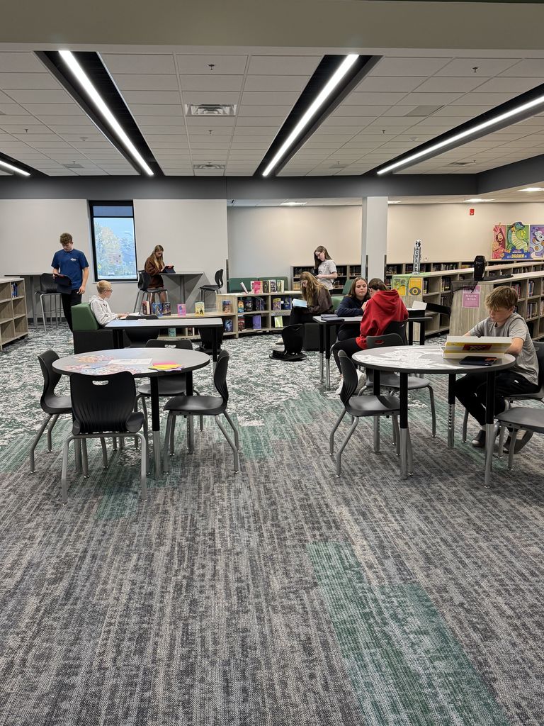 Students in a library working at tables. Two in the back are looking at new books. Two are also working on their iPads. 