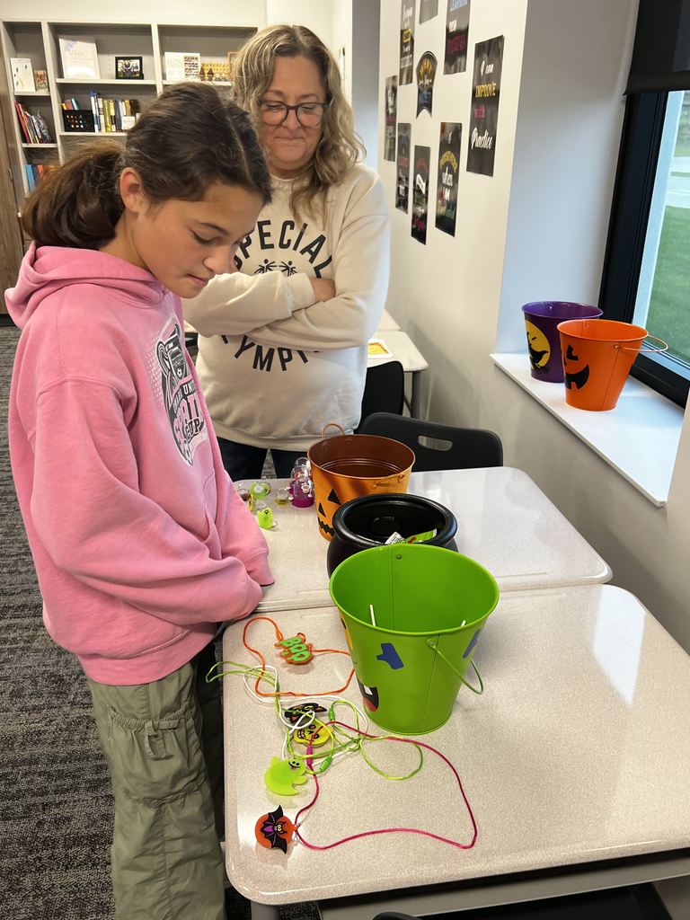 A student stands in front of a desk to play a Halloween game. An adult woman stands besides her. 