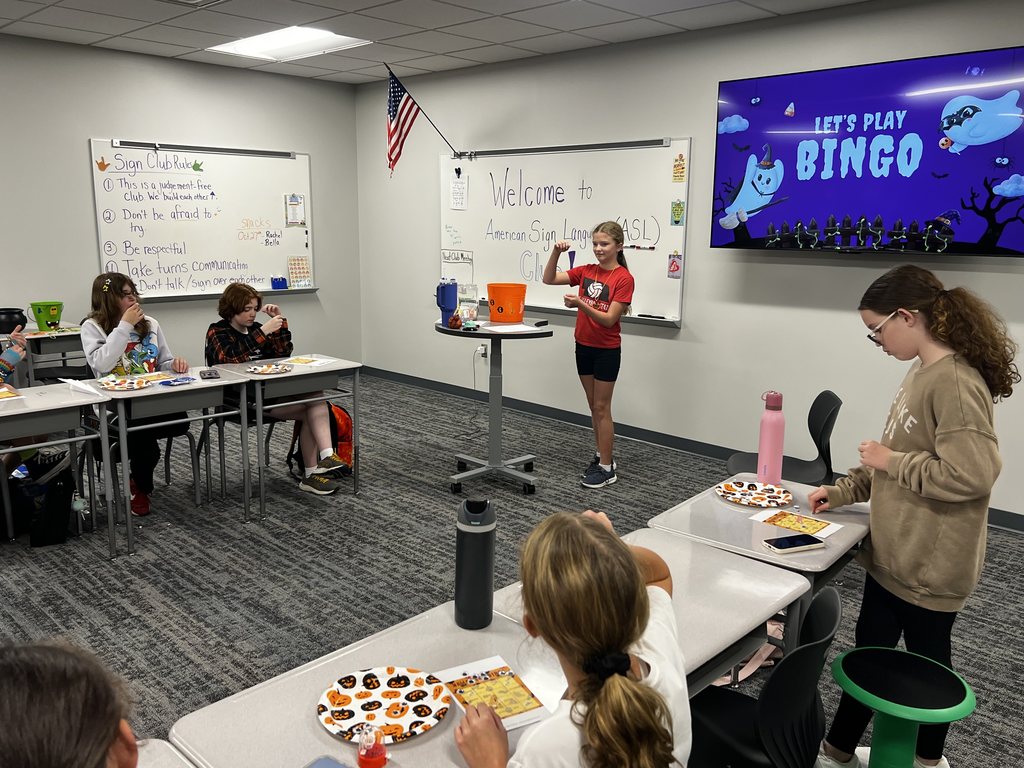 A girl stands in front of the class and is signing. There is a group of students at the desks in front of her. They are playing Bingo. 