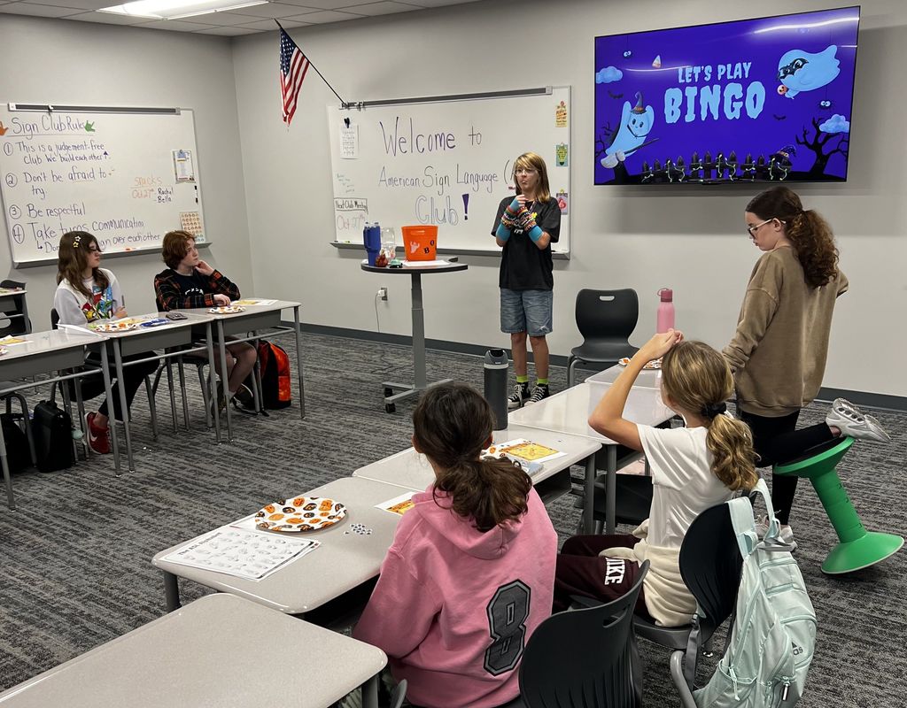 A girl stands in front of the class and is signing. There is a group of students at the desks in front of her. They are playing Bingo. 