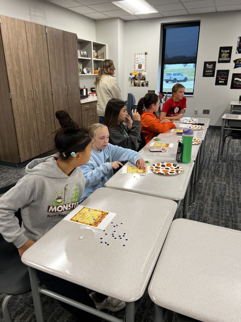 Five students sit at desks playing Bingo with googly eyes as markers. An adult woman stands behind them. 