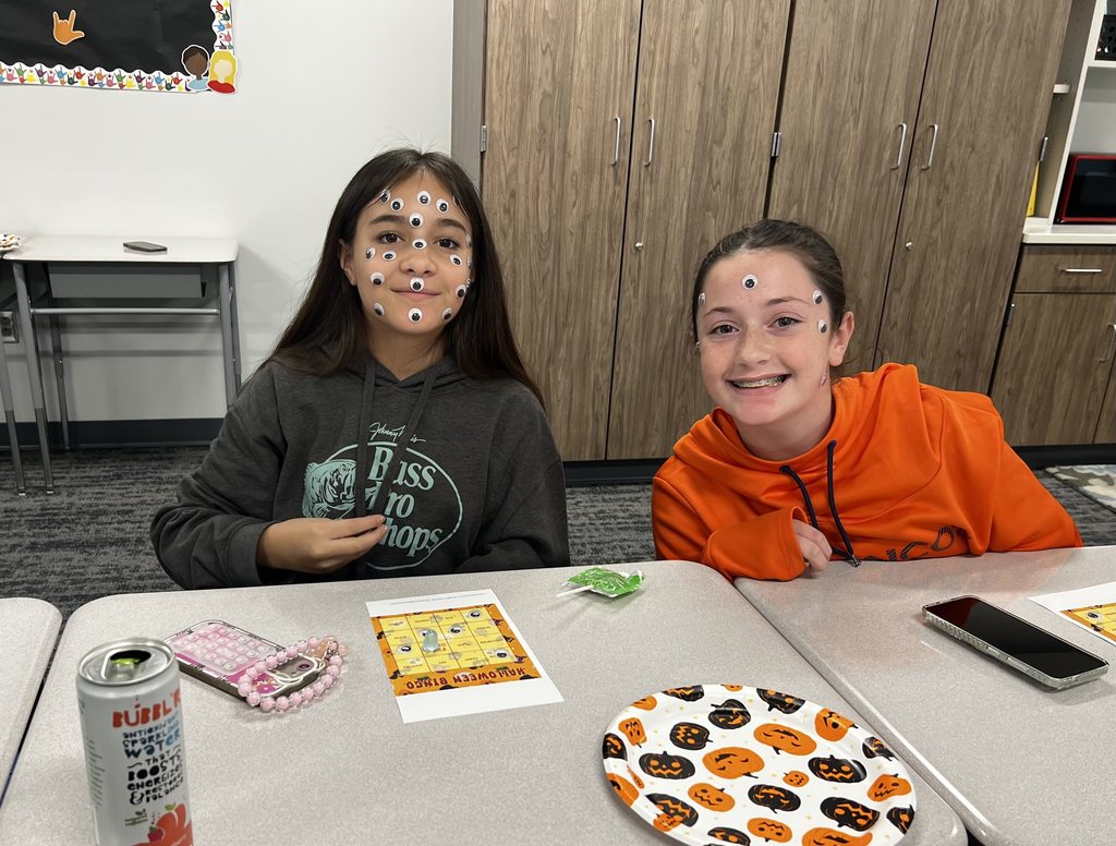 Two girls sitting at desks smile at the camera. They have googly eyes on their faces. 