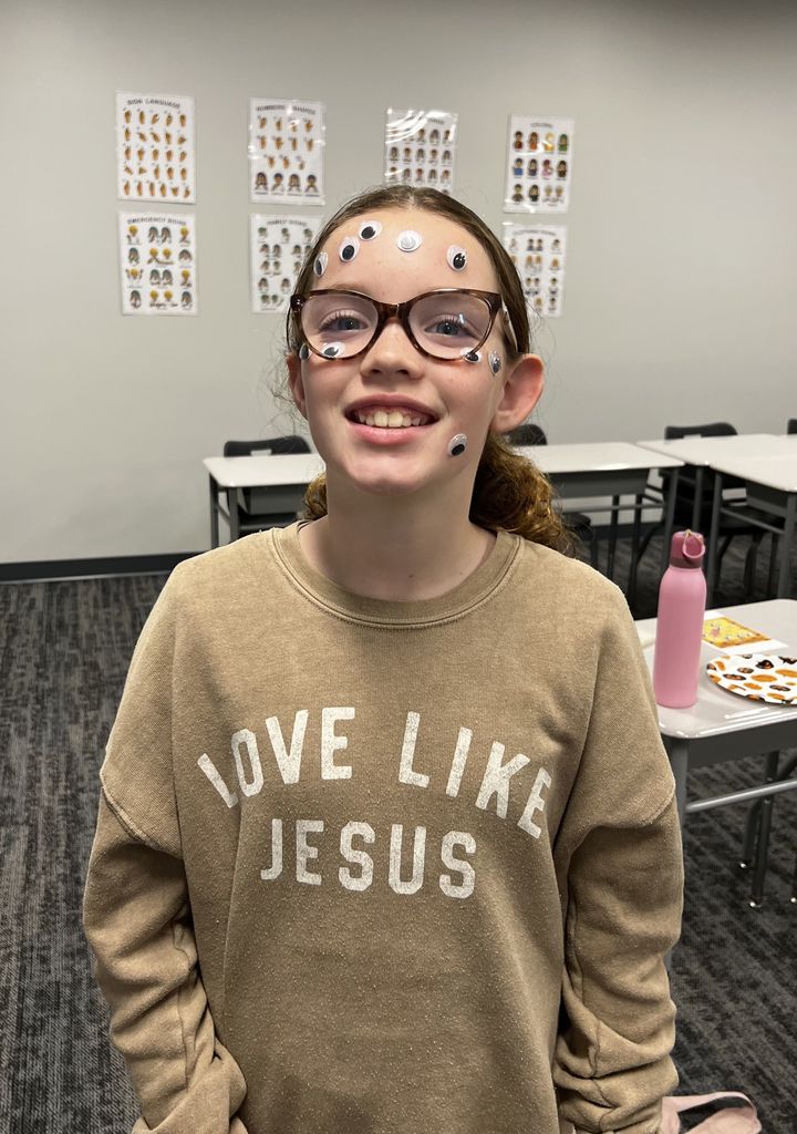 A girl smiles at the camera. She is wearing a "love like Jesus" shirt and has googly eyes on her face. 