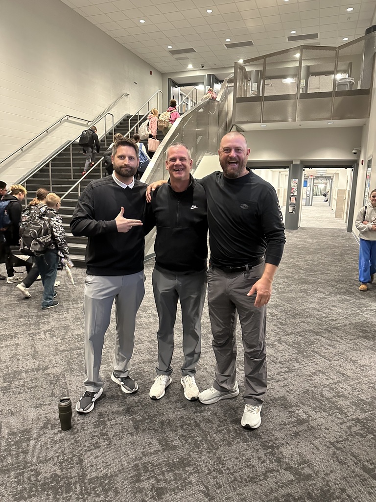 Three adult men stand together with their arms around each other. The one on the left is pointing at the ones on the right. They are all wearing black shirts and gray pants. 