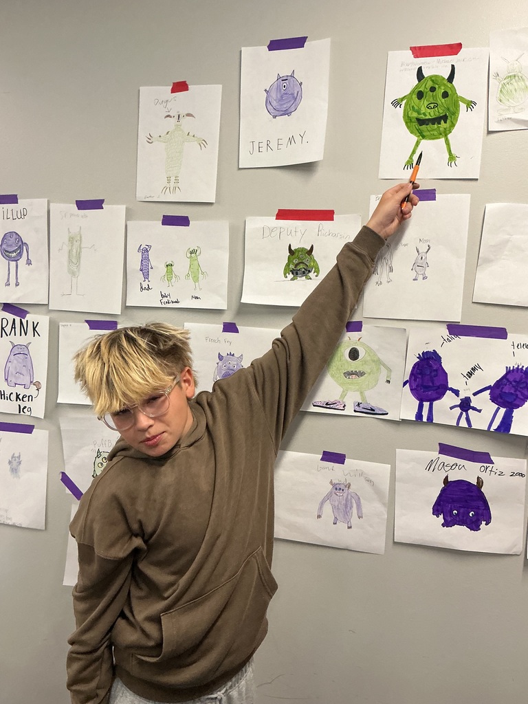 A student stands at the wall, pointing at his green hand drawn monster. 
