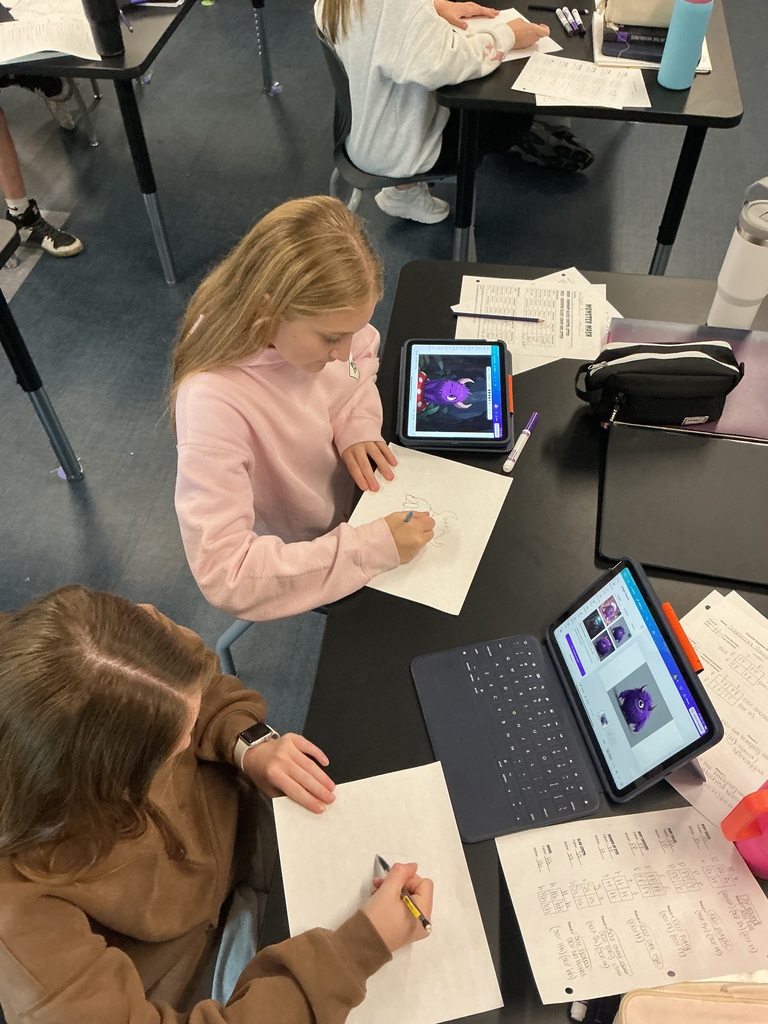 Two students are sitting at a desk working on a piece of paper. There are other papers, ipads, and books on the table. 