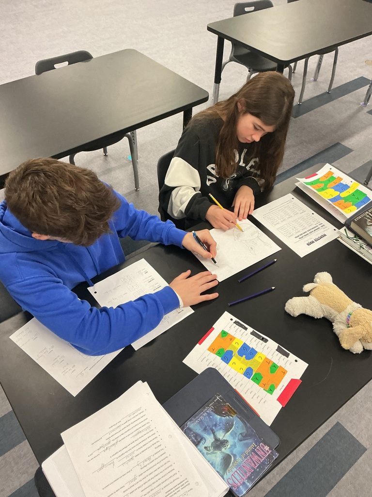 Two students are sitting at a desk working on a piece of paper. There are other papers and books on the table. 