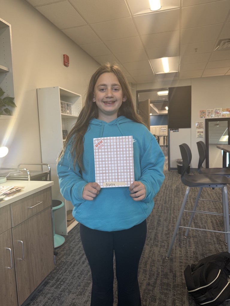 A girl in a blue sweatshirt is holding her pink and white scrapbook page in front of her. She is standing in a classroom. 
