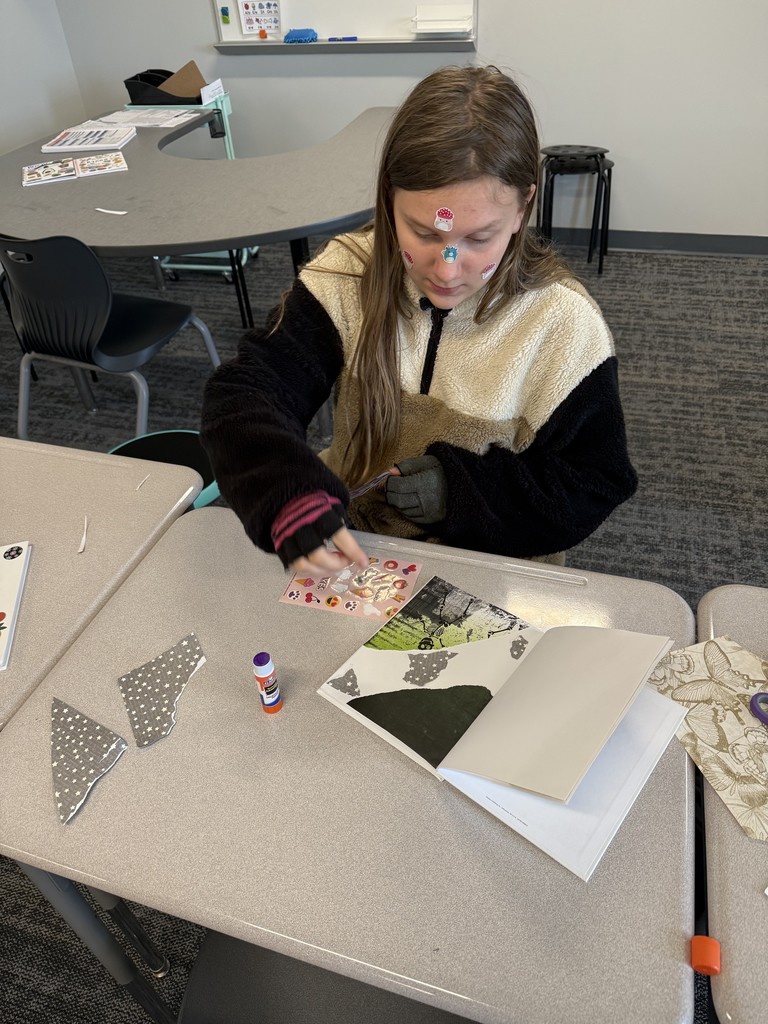 A student is sitting at a desk working  with stickers for her scrapbook page. She has stickers on her face. 