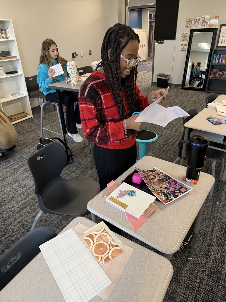 A student is standing up and using scissors to cut out items for her scrapbook page. There is a girl behind her seated at a desk. 