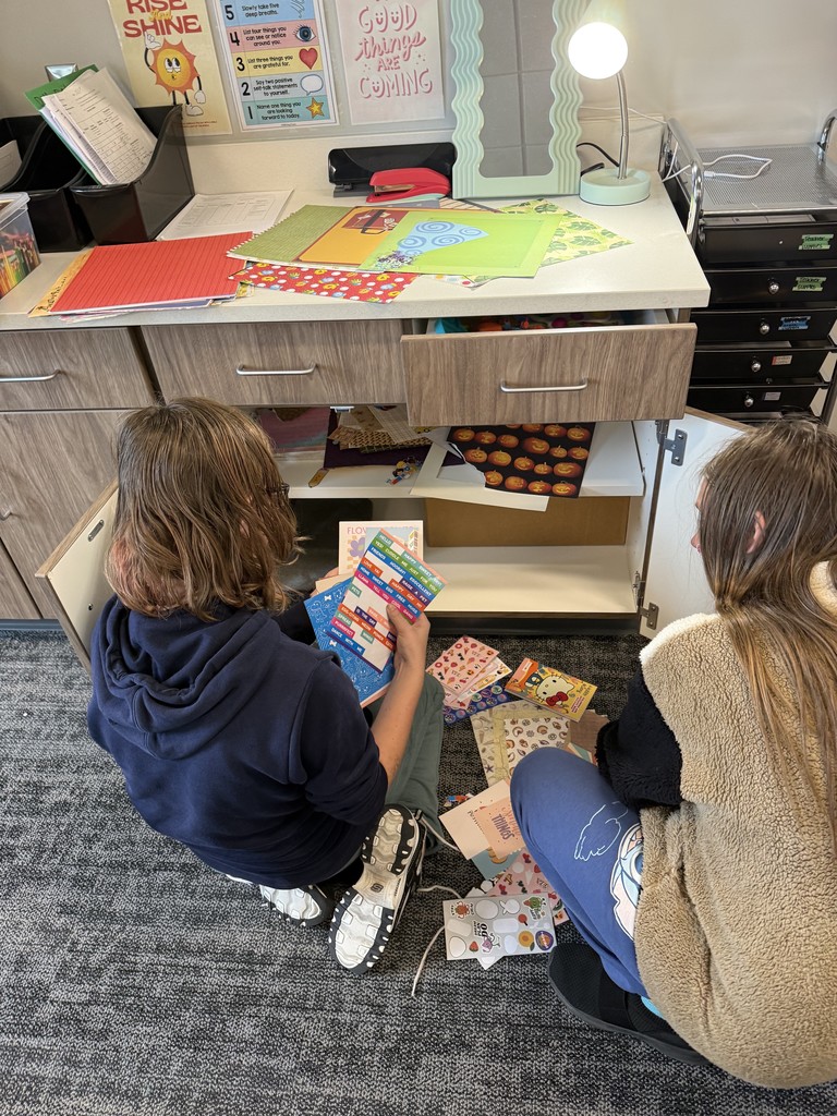 Two students are sitting on the floor with their backs to the camera. They are picking out scrapbook paper from a cupboard underneath a counter. 
