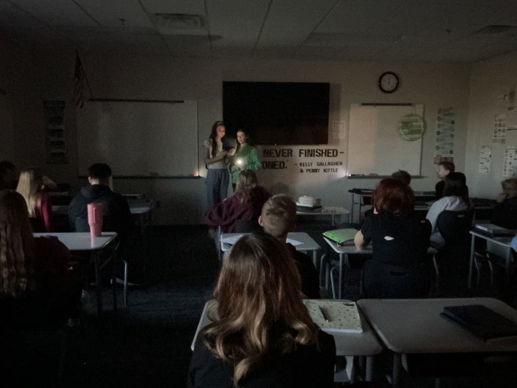 Two students stand in front of the class with a flashlight. The lights are off and they are reading from their story.