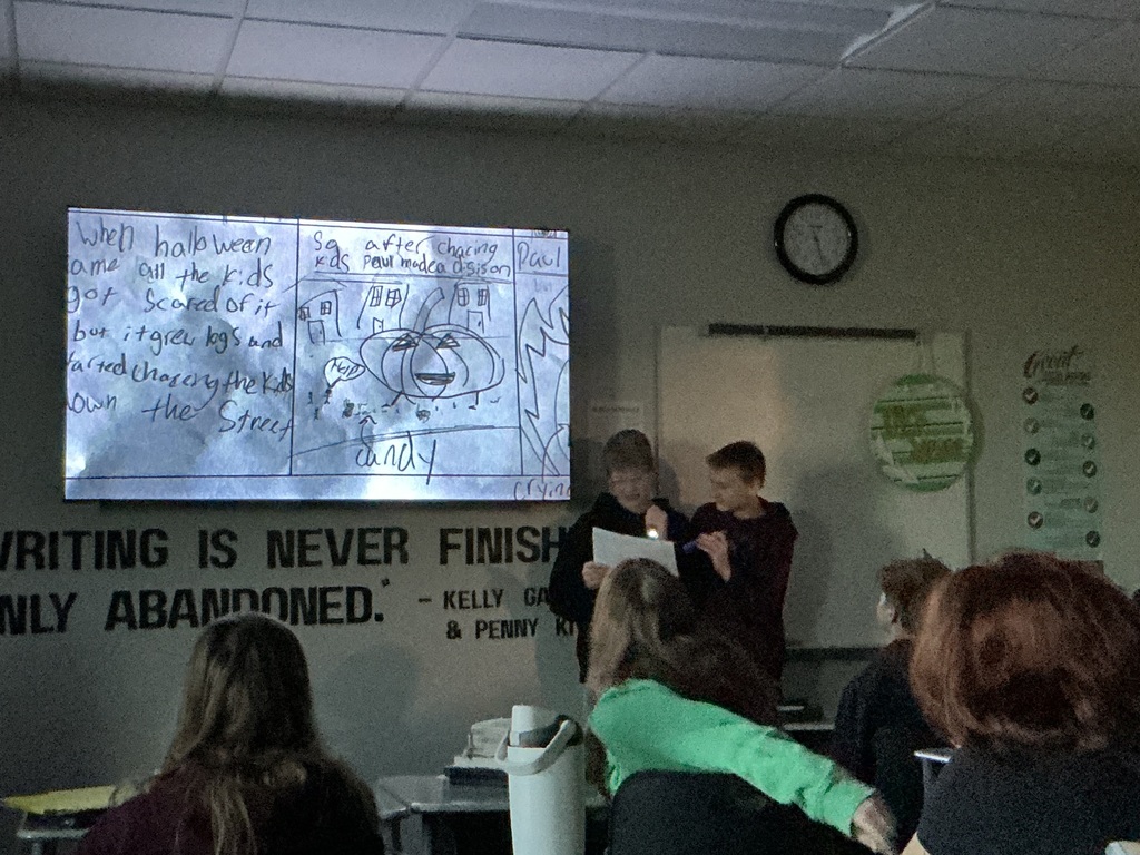 Two students stand in front of the class with a flashlight. The lights are off and they are reading from their story. On the board is their cartoon. 