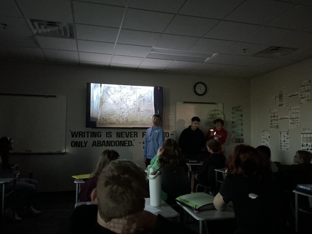 Three students stand in front of the class. The lights are off and they have a flashlight as they read their story. The cartoon is projected on the front TV