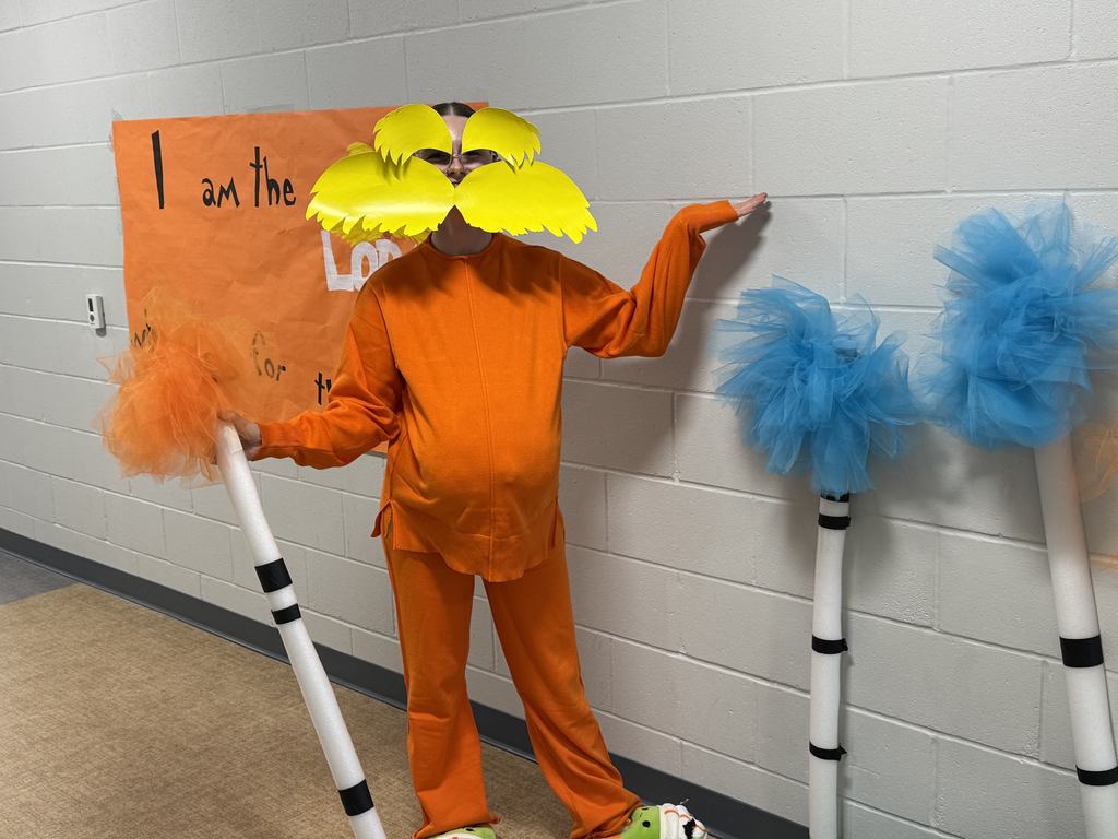 A person is dressed up in orange like the Lorax from the movie and is standing next to decorative fake blue trees. 