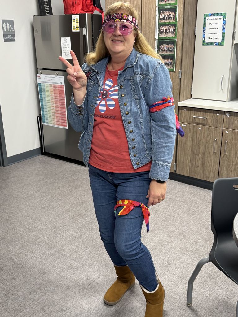 A woman is dressed in 60s/70s garb holding up a peace sign and smiling. 