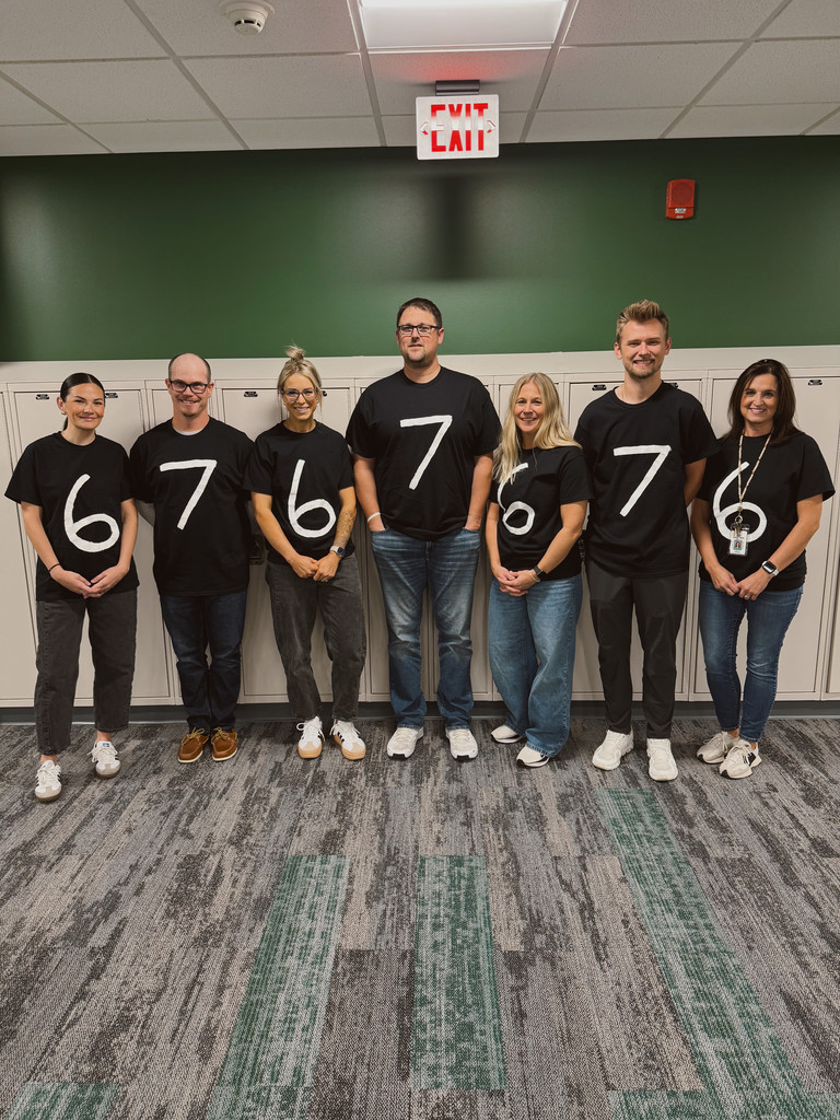 A group of adults is standing in front of lockers. They all have black shirts on. They alternate and one has the number 6 on it, the next has the number 7, and then it repeats. 