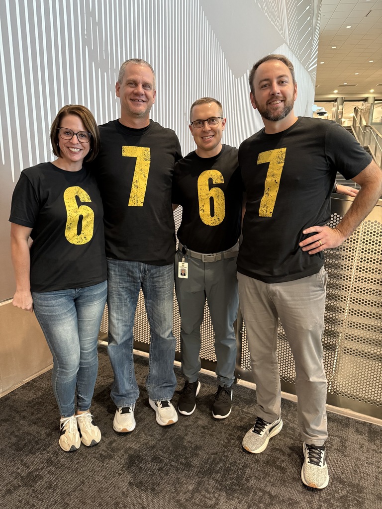 A group of 4 adults is standing together. They have on dark gray shirts that say the number 6 or 7. It alternates. 