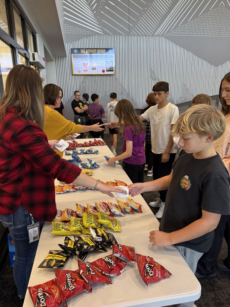 Students are lined up at two long white tables turning in tickets to two women for a chance to pick out an individual bag of chips. 